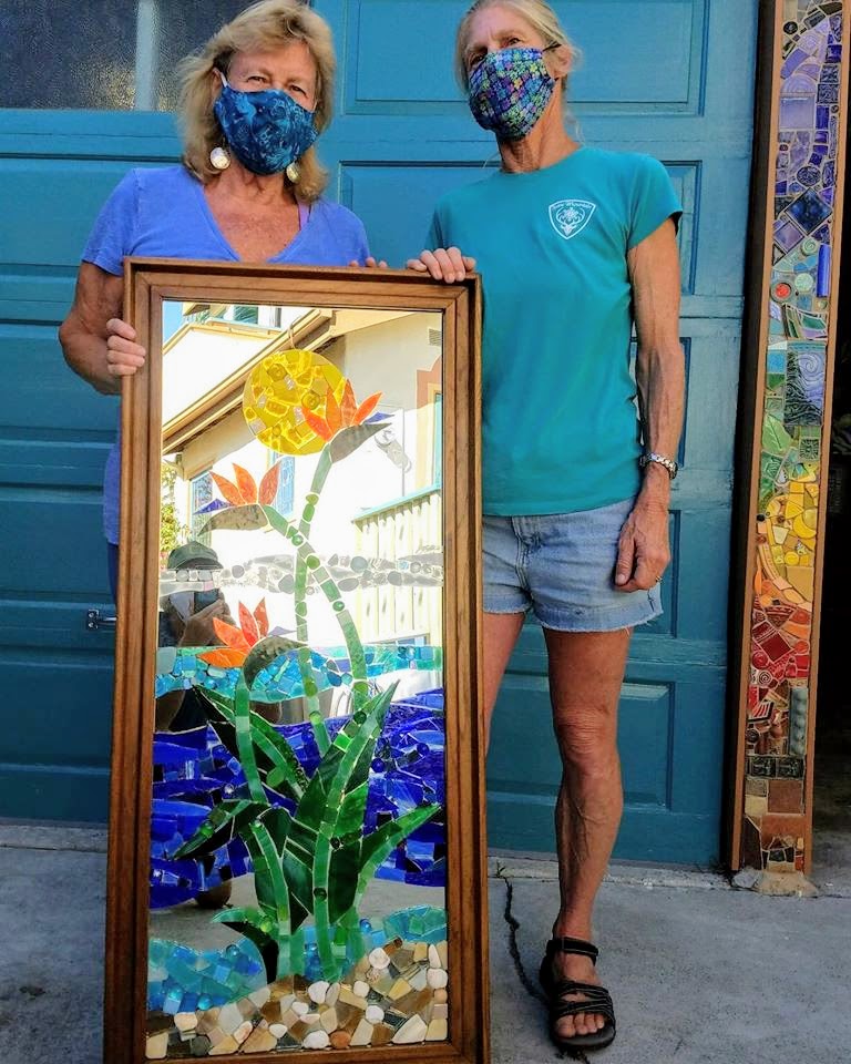 Kathryn standing outside her studio space with a client.  She's holding up the finished mirror.  It has a bird of paradise plant and an ocean motif.